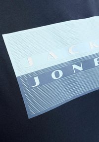 Dunkelblaues T-Shirt mit einem strukturierten hellblauen Motiv, das die Worte "JACK" und "JONES" in fetten Buchstaben auf einem mehrschichtigen Hintergrund zeigt.