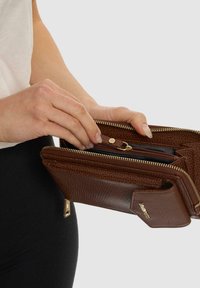 Portafoglio in pelle marrone con finitura testurizzata, zipper dorato e molteplici scomparti. Mano che apre una sezione con zip su uno sfondo chiaro.