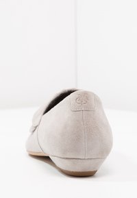 Lichtgrijze suède instapschoen met platte zool, schuin weergegeven om de achterkant en zijkant te tonen, voorzien van een gestikte logopatch op de hiel en een beige zool.