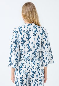 Weißer Stoff mit blauem Blumendruck. Hat drei Viertelärmel, einen lockeren Schnitt und eine taillierte Taille. Weiches, strukturiertes Material.