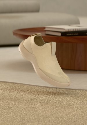 Lys beige slip-on sneaker med teksturert strikket overdel, rund såle og drattab, vist på et teppe nær et trebord.