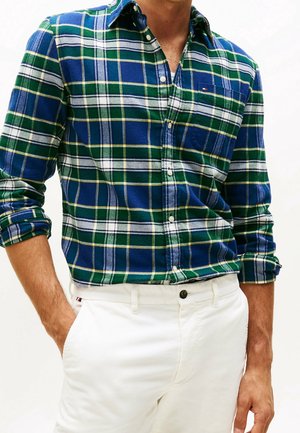 Homme portant une chemise à carreaux bleue, verte et blanche à boutons, manches retroussées, et un pantalon blanc avec une main dans la poche.