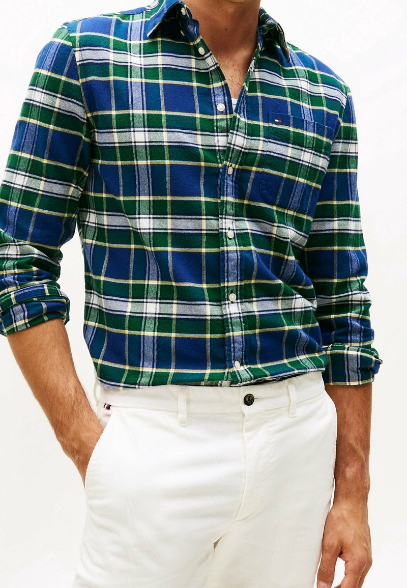 Homme portant une chemise à carreaux bleue, verte et blanche à boutons, manches retroussées, et un pantalon blanc avec une main dans la poche.
