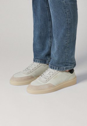 Weiße und beige flache Sneaker mit Schnürsenkeln, getragen mit blauen Jeans vor einem schlichten Hintergrund.
