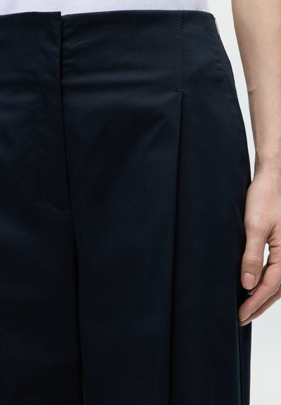 Gros plan sur la main d'une personne reposant à côté d'une jupe plissée noire et d'un haut blanc, mettant en valeur la texture du tissu et les détails du vêtement.