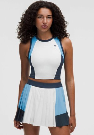 Femme aux cheveux bouclés portant un crop top blanc sans manches et une jupe plissée avec des blocs de couleurs blanc, bleu clair et bleu foncé sur un fond uni.
