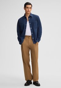 Donkerdenim overhemd met knopen over een witte tanktop, gecombineerd met beige broek en zwarte veterschoenen. Het overhemd heeft twee borstzakken.