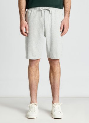 Homme portant un short gris clair à cordon de serrage et des baskets blanches, debout devant un fond uni.