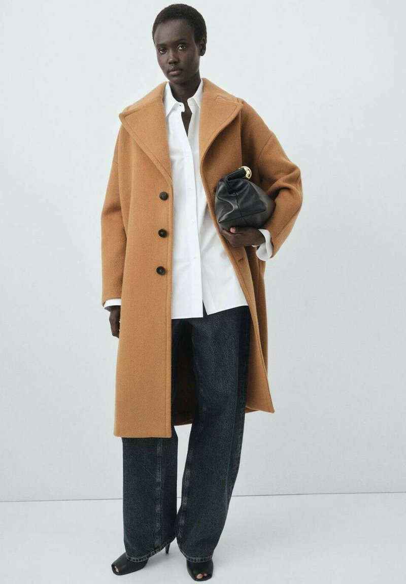 Abrigo de lana color camel con grandes solapas, botones negros, combinado con una camisa blanca, jeans oscuros de pierna ancha y un bolso de cuero negro.