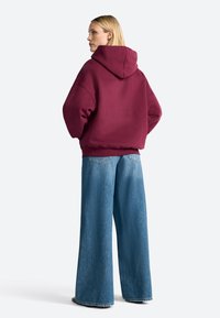 Bordeaux hoodie met een losse fit en geribbelde manchetten. Gecombineerd met lichtblauwe denim jeans met wijde pijpen, voorzien van achterzakken en een vervaagde textuur.