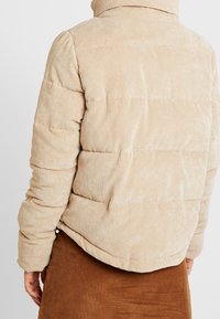 Beige kordfløj pufferjakke med høj krave og quiltet design. Har ribbet tekstur og en let oversized pasform.