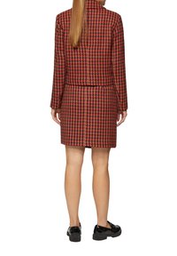 Czerwono-czarny żakiet w kratę houndstooth w połączeniu z dopasowaną spódnicą, obie części o teksturowanym wykończeniu. Modelka nosząca czarne buty sznurowane.