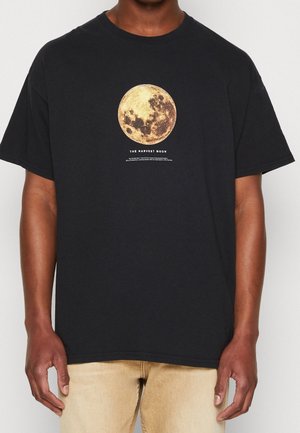 Personne portant un T-shirt noir avec un grand motif de pleine lune jaune et le texte "The Harvest Moon" sur la poitrine, associé à un pantalon marron clair.
