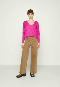 Suéter rosa brilhante, largo, com decote em V, combinado com calças largas bejes e sapatos de plataforma castanhos. O modelo usa um boné castanho.