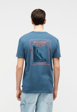Personne portant un t-shirt Billabong bleu avec un graphisme de surf orange et du texte au dos, associé à un jean en denim bleu clair.