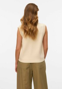 Top en tricot sans manches beige avec un col et un ourlet côtelés, associé à un pantalon large olive avec des poches arrière doubles.