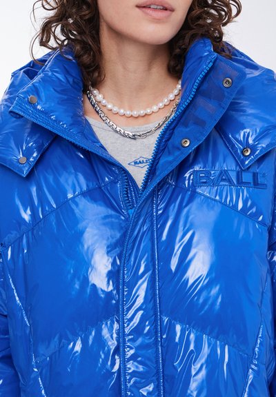 Mėlynas pūkinis džemperis su blizgiu paviršiumi, turintis užtrauktuką priekyje, aukštą apykaklę ir trikampį siuvinėjimo raštą. Įtraukti siuvinėti logotipo elementai.