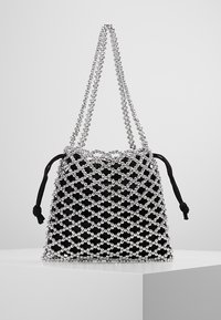 Sac en tissu noir avec un revêtement en filet de perles argentées et une double bandoulière en perles argentées, posé sur un piédestal blanc devant un fond uni.