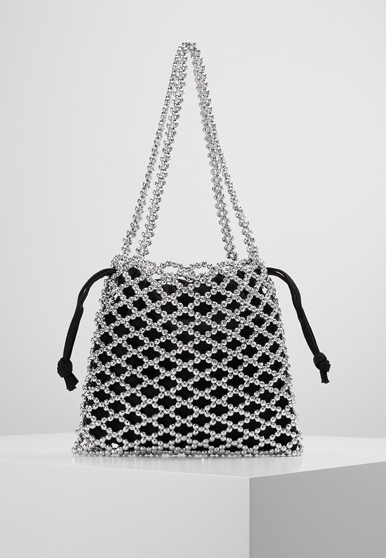 Sac en tissu noir avec un revêtement en filet de perles argentées et une double bandoulière en perles argentées, posé sur un piédestal blanc devant un fond uni.