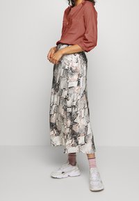 Blouse couleur rouille avec col en V et manches retroussées, assortie à une longue jupe à motifs dans des tons de noir, blanc et beige. Baskets blanches.