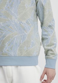 Lyseblå sweatshirt med et bladrigt grønt og hvidt mønster. Har ribbede mansjetter og kant i ensartet lyseblå. Blød tekstur.