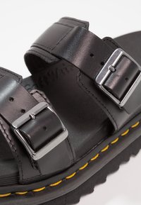 Sandal in pelle nera con due cinturini regolabili, fibbie in argento e una suola testurizzata. Presenta un dettaglio di cucitura gialla sulla suola.