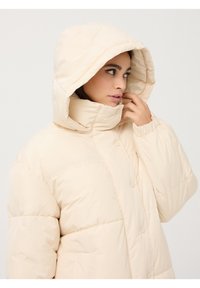 Beżowa kurtka puchowa z dużym kapturem, zatrzaskami i pikowanym wzorem. Posiada wysoki kołnierz i boczne kieszenie. Odpowiednia na zimną pogodę.