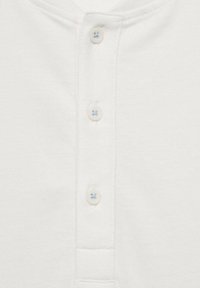Vit kortärmad henley-tröja med fyra runda knappar med ljusblå accenter, en ribbad krage och en slät tygtextur.