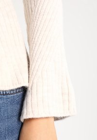 Licht crèmekleurig geribbeld sweater met flared mouwen, zachte textuur en een ronde zoom. Gecombineerd met blauwe spijkerbroek bij de zoom.