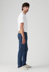 Modri jeans s ravnim krojem, opremljeni z usnjeno nalepko na hrbtni elastiki; v kompletu z navadno belo majico s kratkimi rokavi.