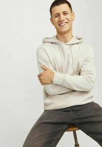 Sudadera beige claro con cordones, tela suave y logo en la parte frontal; combinada con jeans gris oscuro, sentada en un taburete de madera.