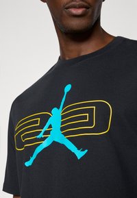 Czarna bawełniana koszulka z dużą niebieską sylwetką skaczącej postaci oraz żółtym napisem, okrągłym dekoltem i krótkimi rękawami.