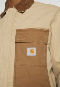 Beige en bruine jas met een grote voorzak, voorzien van een geel Carhartt logo patch en een corduroy kraag.