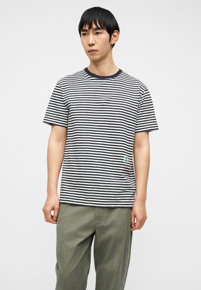 Άνδρας που φοράει μαύρο και λευκό οριζόντια ριγέ μπλουζάκι και χακί παντελόνι, στέκεται μπροστά σε απλό λευκό φόντο.