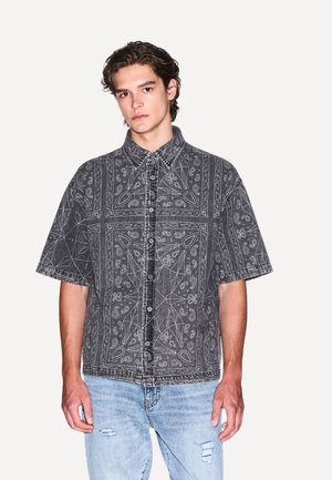Camicia a maniche corte con bottoni in denim nero, caratterizzata da motivi paisley bianchi e colletto. Indossata con jeans strappati blu chiaro.
