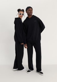 Des sweats à capuche et des pantalons noirs en tissu doux, avec une coupe décontractée. Un modèle porte des lunettes de soleil blanches et l'autre un bonnet noir.