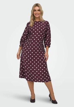 Donna che indossa un vestito midi viola con pois rosa e maniche a sbuffo a tre quarti, in piedi con una gamba leggermente avanti, sorridente.