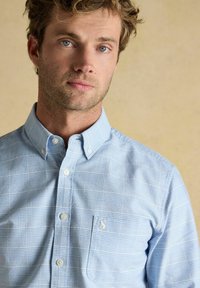 Detailný záber na muža so svetlo hnedými vlasmi, modrými očami a strniskom, ktorý má na sebe svetlomodrú kockovanú košeľu s vreckom na hrudi.