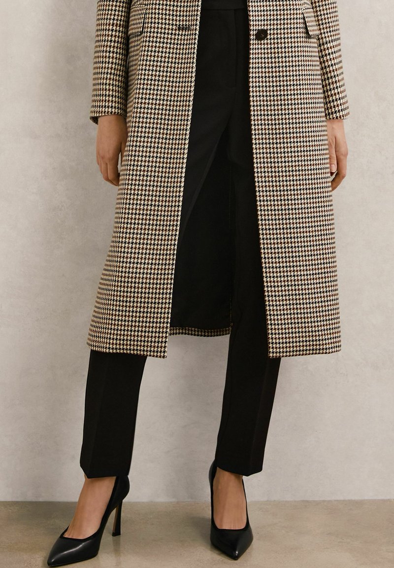 Manteau à motif pied-de-poule marron et beige avec une coupe cintrée, associé à un pantalon noir et des chaussures à talons noires. Textures lisses dans l'ensemble.