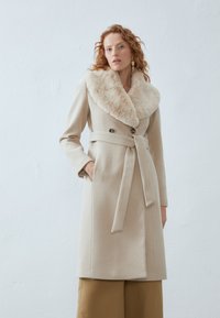 Beige wool coat with a textured faux fur collar, double-breasted buttons, and a tied waist belt. Smooth fabric and tailored fit.
