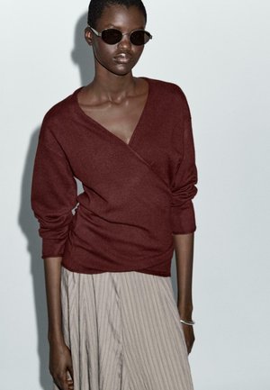 Cardigan bordeaux avvolgente in morbido mix di lana con maniche lunghe, abbinato a una gonna plissettata di un leggero beige. Modella che indossa occhiali da sole scuri.
