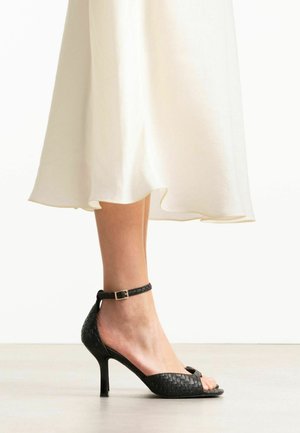 Femme portant des sandales noires tissées à talons hauts avec bride à la cheville et une jupe midi blanche, debout sur un sol clair.