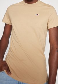 Béžové bavlněné tričko s kulatým výstřihem, krátkými rukávy a malým vyšitým logem v horním levém rohu, s hladkým povrchem.