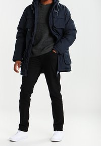 Námořnický pufovaný kabát s kapucí na stahování, několik předních kapes, šedý pletený svetr, černé slim-fit kalhoty a bílé tenisky.