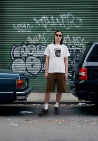 Vit t-shirt med svart "FORMER"-tryck, bruna cargobyxor, svarta skor, vita strumpor. Omgiven av parkerade bilar och en bakgrund med graffiti.