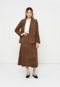 Blazer en daim marron avec un bouton unique, associé à une jupe assortie jusqu'aux genoux. Le modèle porte un haut court blanc et des ballerines noires.