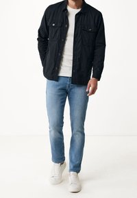 Black button-up shirt with two chest pockets, worn over a white tee, paired with light blue jeans and white sneakers. Smooth fabric texture.