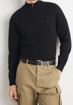 Homme portant un pull noir à fermeture éclair quart de zip avec un logo "BOSS" subtil, associé à un pantalon cargo beige et une ceinture en cuir noire avec une boucle argentée.