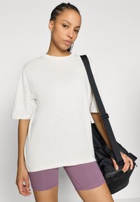 Φαρδιά ανοιχτό μπεζ μπλούζα με κοντά μανίκια, σε συνδυασμό με εφαρμοστά μωβ σορτς. Το μοντέλο κρατά μαύρη τσάντα γυμν αστηρίου με ιμάντα ώμου.