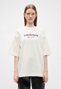 Λευκό oversized t-shirt από βαμβάκι, με λογότυπο "CALVIN KLEIN NEW YORK" σε μαύρο και κοραλί, κοντά μανίκια και άνετη εφαρμογή.